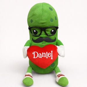 May include: A plush green pickle toy with black glasses and a mustache, holding a red heart with the name "Daniel" in white. The pickle has white gloves and striped socks. A whimsical and fun gift.