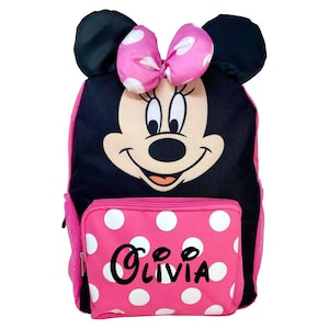 May include: A pink and black backpack featuring a Minnie Mouse design. The backpack has a pink polka-dot front pocket with the name "Olivia" in black script. The backpack includes black mouse ears and a pink bow with white polka dots.