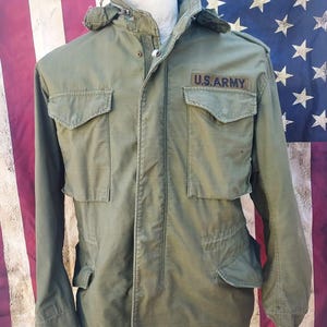 Può includere: Giacca militare verde oliva con più tasche e cappuccio. La giacca presenta una toppa "U.S. ARMY" sul petto. La giacca è esposta su uno sfondo di una bandiera americana.