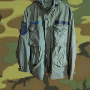 May include: An olive green military jacket with multiple pockets, a hood, and the text "AIR FORCE" on the chest. The jacket also features a patch with a blue and white emblem and the name "SPADORA".