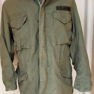 Puede incluir: Una chaqueta de campo verde oliva con cuello alto y capucha, cierre frontal de cremallera y cuatro bolsillos grandes con solapa y botón. Un parche negro con el texto U.S. ARMY está cosido sobre el bolsillo del pecho izquierdo.