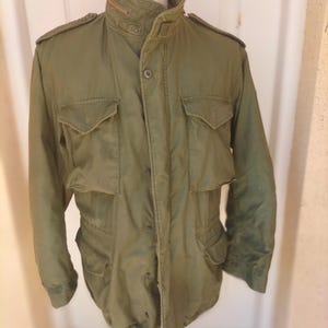 May include: An olive green field jacket featuring a high collar with a concealed zipper, shoulder epaulets, and four large front pockets with buttoned flaps. The jacket includes a central button closure and a structured, durable fabric construction.