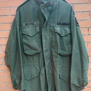 May include: An olive green military jacket with multiple pockets and a button-up front. The jacket has the text "MEDINA" above the left pocket and "U.S. ARMY" above the right pocket. A classic piece of military apparel.