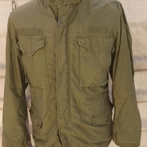 May include: An olive green field jacket featuring a high collar with a zipper, a front button closure, and four large flap pockets on the chest and waist. The jacket includes long sleeves and a structured design, displayed on a mannequin torso.