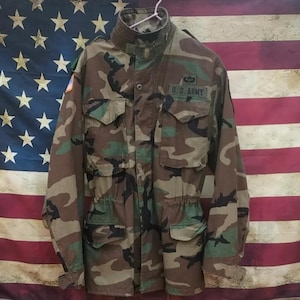 Può includere: Giacca militare mimetica con più tasche e la scritta "U.S. ARMY" su una toppa. La giacca ha una chiusura con bottoni, un colletto alto e una vita con coulisse. Lo sfondo è una bandiera americana.