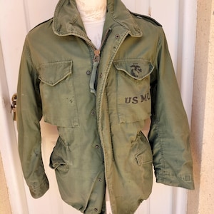 Puede incluir: Una chaqueta de campo verde oliva con cuello alto, cierre de cremallera frontal y cuatro bolsillos delanteros. El bolsillo izquierdo del pecho muestra un emblema USMC negro y el texto US MC. La chaqueta tiene mangas largas y un diseño utilitario.