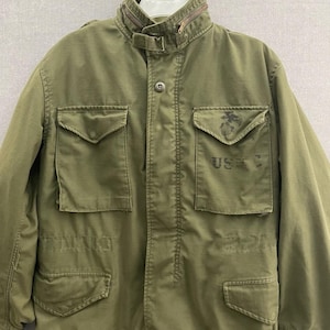 May include: An olive green field jacket featuring a high collar with a zipper, a button-front closure, and four front pockets with buttoned flaps. The right chest pocket displays a faded black USMC emblem and the text USMC printed in black ink.