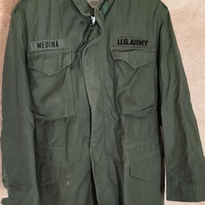 May include: An olive green field jacket featuring a high collar, a front zipper closure, and four large flap pockets. The chest displays black embroidered text reading MEDINA on the left and U.S. ARMY on the right side of the garment.