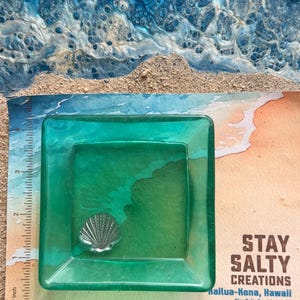 May include: A square, translucent green resin dish featuring a small, silver-colored metal seashell embedded in the center. A ruler placed along the left and bottom edges shows the dish measures approximately 4 inches by 4 inches in size.