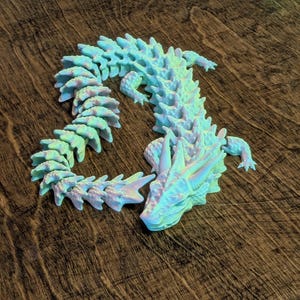 May include: A 3D printed articulated dragon figurine featuring a pearlescent finish that shifts between light blue, green, and soft pink hues. The creature has a segmented, flexible body with sharp dorsal spikes and a detailed head with horns.
