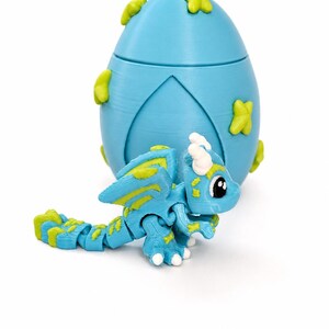 May include: A blue articulated dragon toy with yellow accents and a matching blue egg-shaped container. The dragon has a detailed design with wings and a long tail. The egg has yellow leaf-shaped decorations.