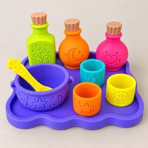 May include: A set of colorful silicone potion-making toys including a purple tray, a purple cauldron with a yellow spoon, three bottles in lime green, orange, and pink with tan stoppers, and three small cups in teal, orange, and yellow with embossed icons.