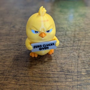 May include: A small yellow chick figurine with an angry expression, featuring blue eyes and an orange beak. The chick holds a rectangular gray sign in its wings that has the black text "ZERO CLUCKS GIVEN" printed clearly on the front surface.