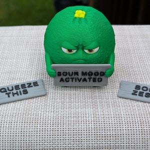 May include: A green, cartoon-like lime character with an angry expression, holding a sign that reads "SOUR MOOD ACTIVATED." Two gray signs nearby say "SQUEEZE THIS" and "BORN ZESTY."