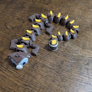 May include: A brown articulated monkey-like figure with a light gray face and large eyes. The figure has a segmented body with small yellow bananas attached to each section. A separate small brown barrel with yellow bananas sits nearby.