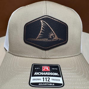 Op de afbeelding: Beige truckerpet met een witte mesh achterkant en een zwart en koperkleurig embleem met een vinontwerp. De pet heeft een gebogen klep en een verstelbare band. Het label van het merk Richardson is zichtbaar.