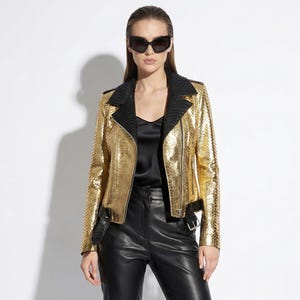 Könnte beinhalten: Eine Frau trägt eine goldfarbene Bikerjacke mit schwarzen Akzenten, ein schwarzes Oberteil und eine schwarze Lederhose. Die Jacke hat ein strukturiertes, schlangenlederartiges Muster. Sie trägt auch eine schwarze Sonnenbrille und Stiefel.