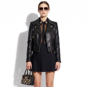 Peut inclure: Une personne portant une veste en cuir noire avec des fermetures éclair dorées, une jupe plissée noire et des bottes noires montantes. Elle porte également des lunettes de soleil et tient un petit sac à main noir avec des ornements dorés et perles.