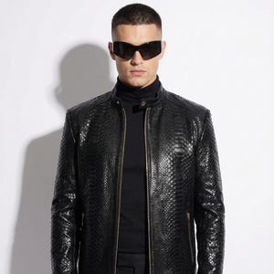 Può includere: Giacca in pelle nera con texture a pelle di serpente, chiusura con zip e collo alto. Indossata con un dolcevita nero e occhiali da sole. Lo stile è elegante e moderno.