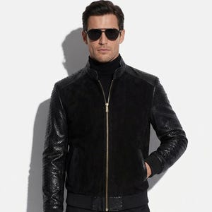 May include: A black jacket with textured leather sleeves and shoulders. The jacket features a high collar, a gold zip, and a ribbed hem. The person in the image is wearing black sunglasses and a black roll neck.