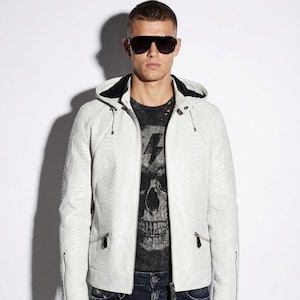 Puede incluir: Chaqueta de cuero blanco texturizado con capucha y cremalleras. La chaqueta se lleva sobre una camiseta negra con gráfico y vaqueros azul oscuro. Las gafas de sol son negras.