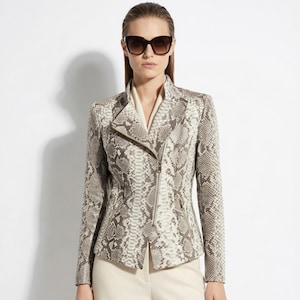 Puede incluir: Chaqueta con estampado de piel de serpiente con cremallera diagonal y cuello alto. La chaqueta es en tonos grises y crema, combinada con pantalones color crema. La modelo lleva gafas de sol.