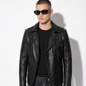 May include: Black leather jacket with a textured collar and shoulder detailing. The jacket has a zipper closure and multiple pockets. The person is wearing black sunglasses, a black t-shirt, and distressed black jeans. A studded belt completes the look.