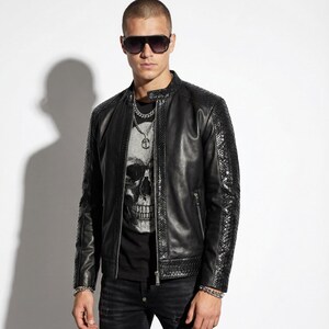 Könnte beinhalten: Schwarze Lederjacke mit strukturierten Ärmeln und Reißverschluss. Das Model trägt ein schwarzes T-Shirt mit Totenkopf-Grafik, schwarze Jeans und Sonnenbrille. Eine silberne Halskette und ein Armband ergänzen den Look.
