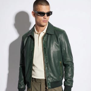 May include: A dark green leather bomber jacket with snakeskin-patterned accents on the shoulders and collar. The jacket has a zip closure and a ribbed hem. The person is wearing a cream-colored polo shirt, olive-green gloves, and sunglasses.