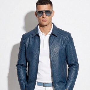 Può includere: Giacca di pelle blu con motivo strutturato, indossata sopra una polo bianca e pantaloni bianchi. La giacca ha una chiusura a zip e una cintura blu abbinata. La persona indossa occhiali da sole.