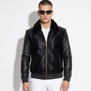 Op de afbeelding: Zwarte bomberjack met een bontkraag en mouwen met slangenleertextuur. De jas heeft een gouden rits en een geribde zoom. De persoon op de foto draagt een zonnebril en een wit overhemd.