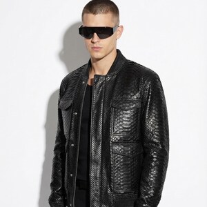 Könnte beinhalten: Schwarze Leder-Bomberjacke mit Schlangenmuster. Die Jacke hat zwei Brusttaschen und einen gerippten Saum und Bündchen. Die Person auf dem Bild trägt eine schwarze Sonnenbrille und eine schwarze Hose.