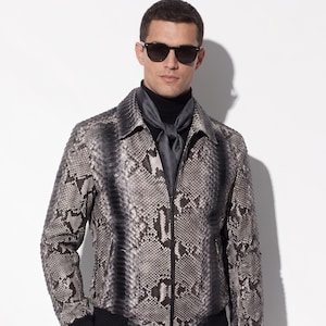 May include: A grey and black snakeskin-patterned jacket with a black collar, cuffs, and hem. The jacket has a zip closure and is worn with a black roll neck and sunglasses. The jacket's design resembles reptile skin.