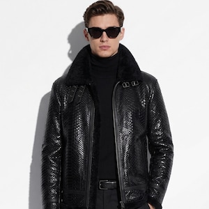 May include: Black leather jacket with a textured pattern, a shearling collar, and a zip-up front. The jacket has a belt and buckle detail at the collar and cuffs. The person in the image is wearing sunglasses and a black turtleneck.