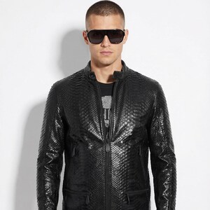 May include: Black leather jacket with a textured, snakeskin-like pattern. The jacket features a zip-up front, a stand-up collar, and multiple pockets. The person in the image is wearing sunglasses and dark-wash jeans.