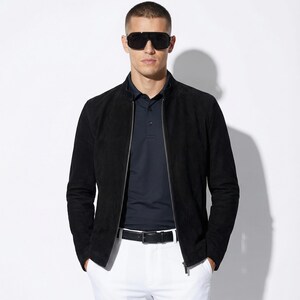 Peut inclure: Veste noire en daim avec fermeture éclair, portée sur un polo bleu marine et un pantalon blanc. L'ensemble comprend une ceinture noire et des lunettes de soleil. Le style est moderne et sophistiqué.