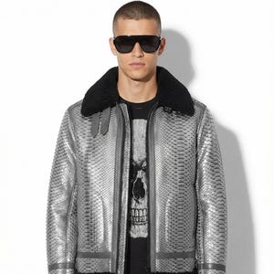 May include: Silver leather jacket with a snakeskin pattern, black shearling collar, and black trim. The jacket has a zip closure and a black t-shirt with a skull graphic underneath. The person is wearing black sunglasses.