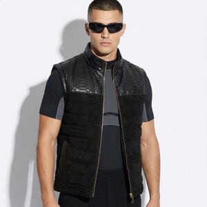 Könnte beinhalten: Schwarze Steppweste mit Schlangenleder-Textur an Schulter und Kragen, mit goldenem Reißverschluss. Die Weste wird mit einem schwarzen T-Shirt und einer schwarzen Hose kombiniert. Der Look ist elegant und modern.