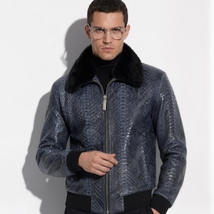 Puede incluir: Chaqueta bomber de cuero azul con estampado de piel de serpiente, cuello de piel negra y dobladillo acanalado negro. La chaqueta tiene cierre de cremallera y se usa sobre un cuello alto negro. El modelo lleva gafas.