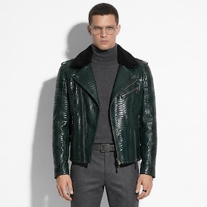 May include: A dark green leather jacket with a textured pattern, black fur collar, and silver zipper. The jacket is paired with a grey turtleneck, grey trousers, a black belt, and black boots. The outfit is completed with eyeglasses.