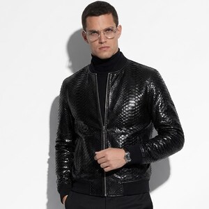 May include: Black leather bomber jacket with a snakeskin texture, featuring a zip-up front and ribbed cuffs. The jacket is paired with a black turtleneck and black pants. A silver watch is visible on the wrist.