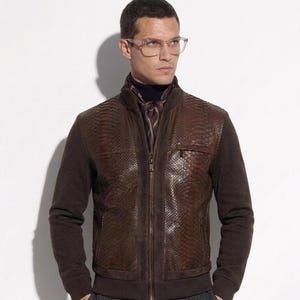 May include: A brown leather jacket with a snakeskin pattern on the front and a zipper closure. The jacket has brown knit sleeves and a high collar. The person is wearing glasses and a patterned scarf. The pants are gray.