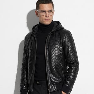 Könnte beinhalten: Schwarze Lederjacke mit Kapuze und strukturierter Schlangenmusterung. Die Jacke hat einen Reißverschluss und gerippte Bündchen. Die Person trägt einen schwarzen Rollkragenpullover und eine schwarze Hose. Eine Brille mit transparentem Rahmen wird getragen.