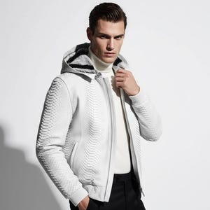 Peut inclure: Veste blanche texturée à capuche et fermeture éclair. Le motif de la veste imite la peau de serpent. Elle est portée avec un col roulé blanc et un pantalon noir. Design moderne et élégant.