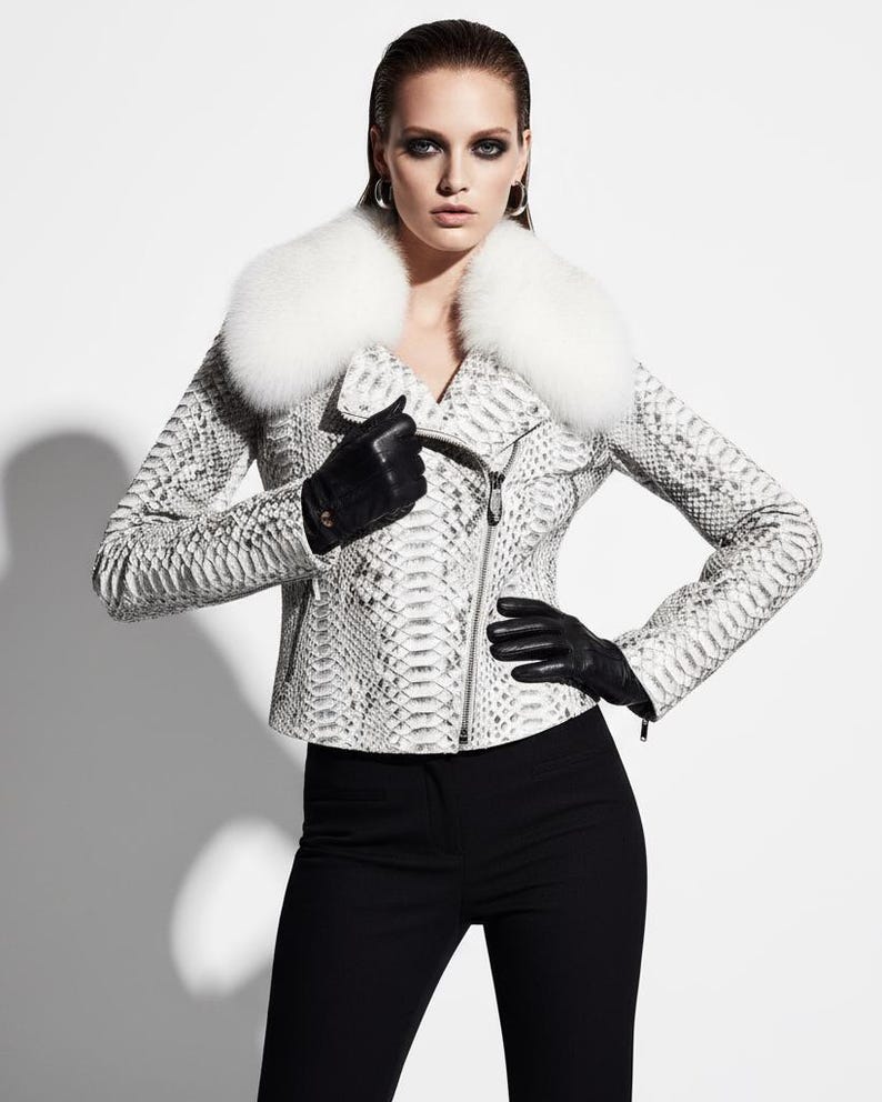 May include: A white and gray snakeskin-patterned leather jacket with a white fur collar. The jacket has a zipper closure and is worn with black gloves and black pants. The model is wearing silver hoop earrings.