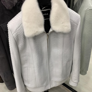 Può includere: Giacca in pelle bianca con motivo a pelle di serpente strutturato. Ha un colletto in pelliccia bianca e una cerniera argentata. Il marchio "ISSLA" è visibile. La giacca è appesa a una gruccia nera. Capo di abbigliamento elegante.