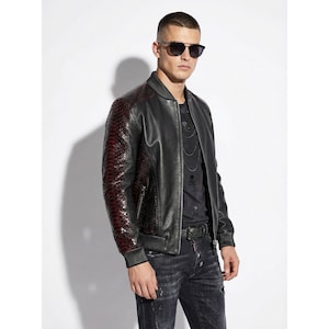 Könnte beinhalten: Schwarze Leder-Bomberjacke mit bordeauxfarbenen Ärmeln mit Schlangenmuster. Die Jacke hat einen gerippten Kragen und Bündchen. Das Model trägt ein schwarzes T-Shirt mit Totenkopf-Design, eine schwarze Jeans und eine Sonnenbrille.