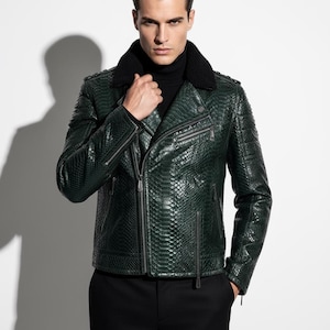 Op de afbeelding: Een donkergroene leren jas met een slangenpatroon, zwarte imitatiebontkraag en meerdere ritsen. De jas wordt gecombineerd met een zwarte coltrui en een zwarte broek, wat een stijlvolle, edgy look creëert.