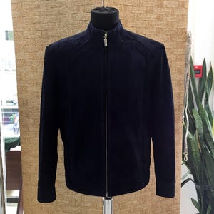 May include: A navy blue suede jacket with a high collar and a full-length zip. The jacket is displayed on a black mannequin head and has a tailored fit. The jacket has a classic design.