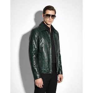 Op de afbeelding: Een donkergroene leren jas met een textuur slangenpatroon. De jas heeft een kraag, een ritssluiting en lange mouwen. De persoon op de foto draagt een zwarte broek en een zonnebril.